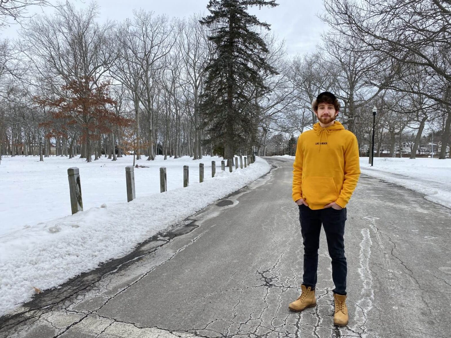 A young man stands in a Portland, Maine park which has snow on the ground. He's wearing a yellow sweatshirt