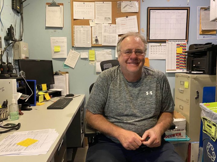 A man in a grey shirt smiles from his chair at a desk