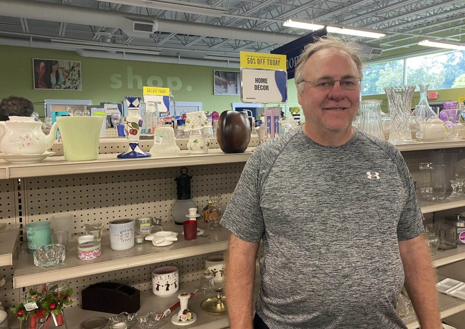 A man in a grey shirt smiles while standing in the glass aisle of a Goodwill store