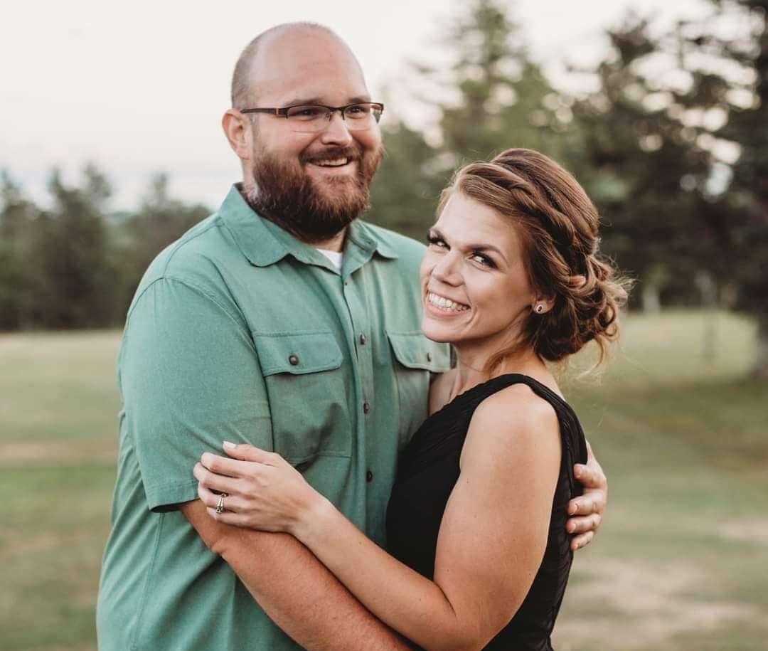 A young woman and a young man hug and look t the camera in front of a green field