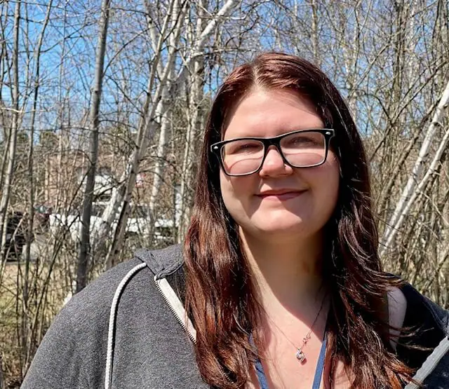 young woman smiles against a wooded background, wearing a sweatshirt