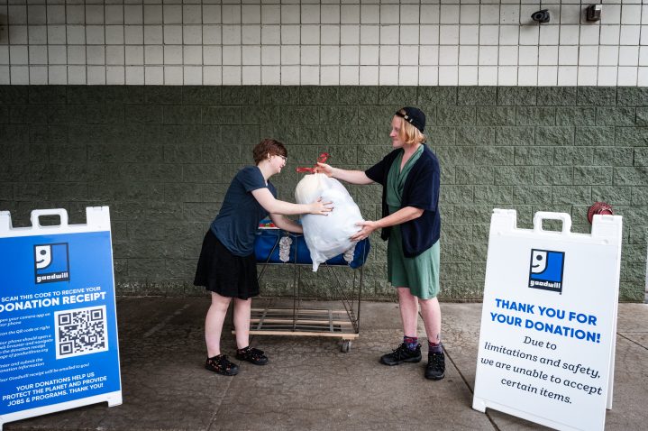 Two individuals exchanging a white trash bag full of donations surrounded by outdoor Goodwill donation signs