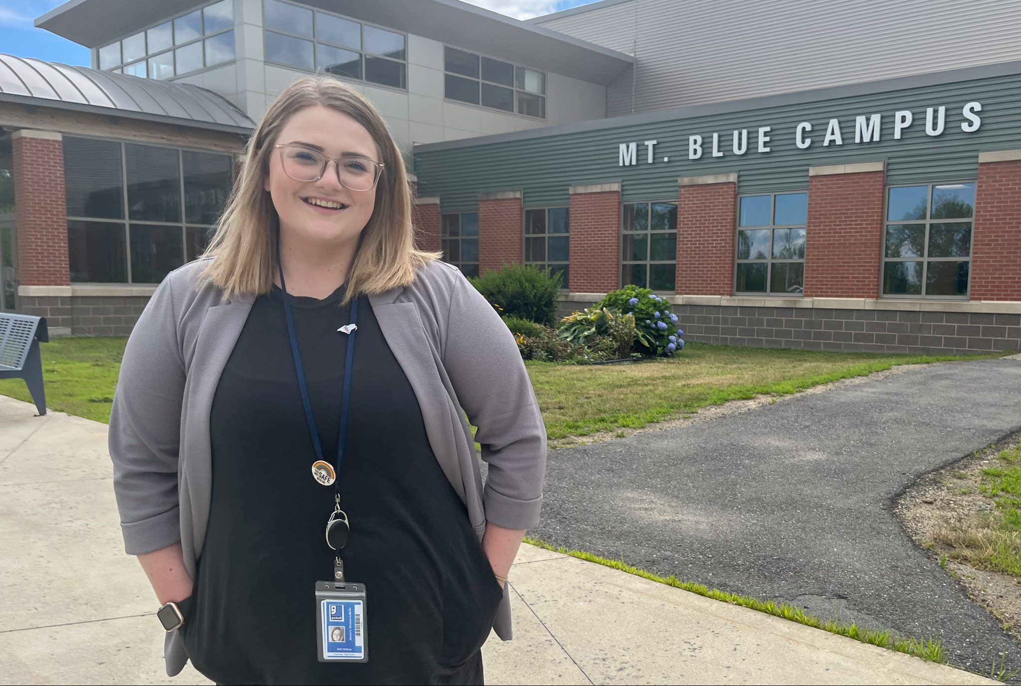 Professional young blonde woman standing and smiling in front of Mt. Blue high school