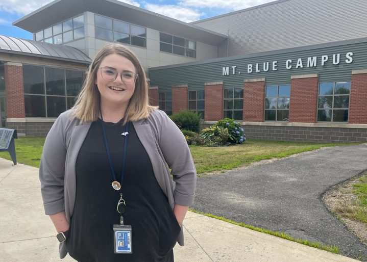 Professional young blonde woman standing and smiling in front of Mt. Blue high school