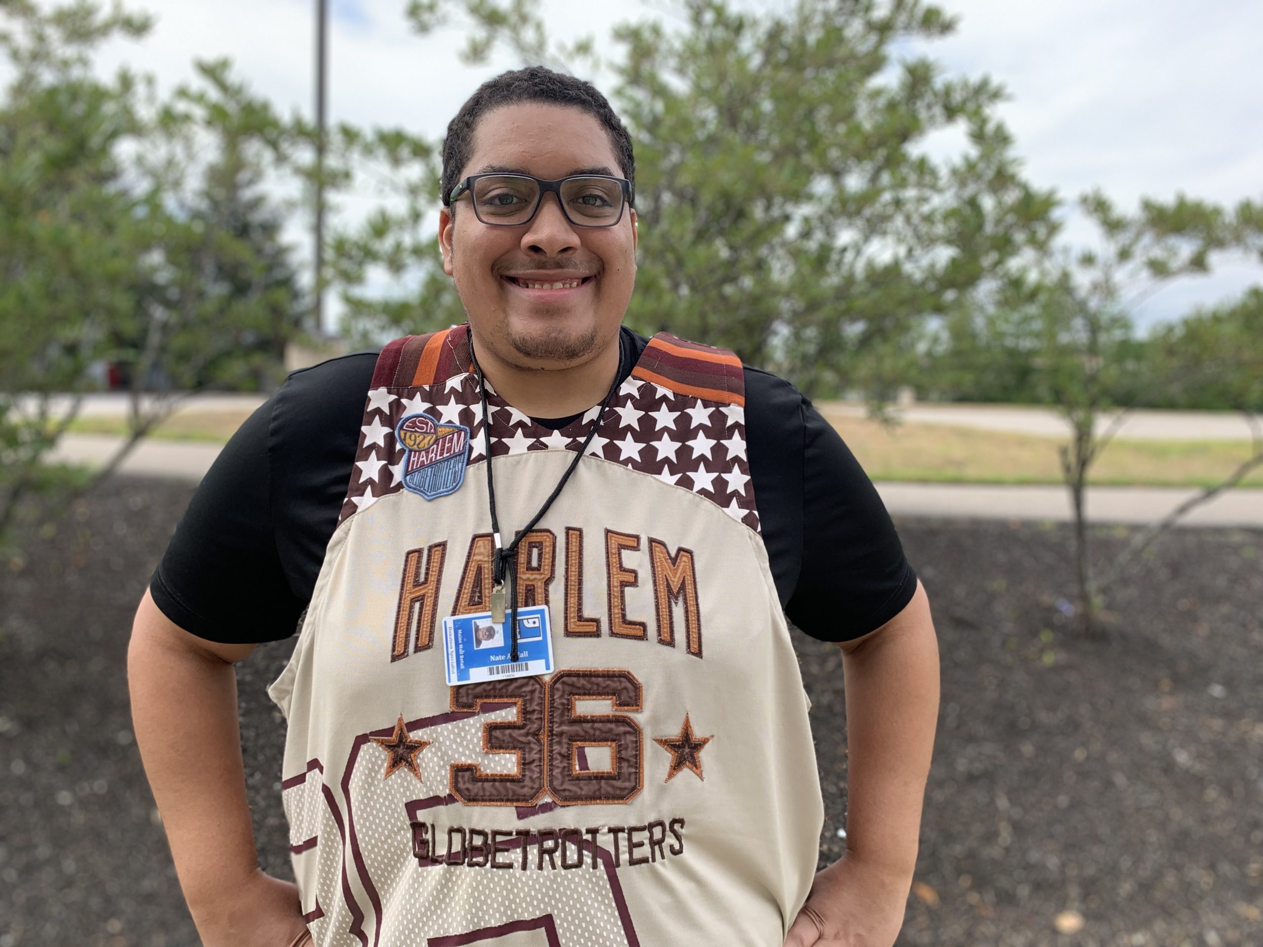 A young man in a Harlem basketball jersey smiles. H'es wearing a Goodwill badge because he's an employee at a donation center