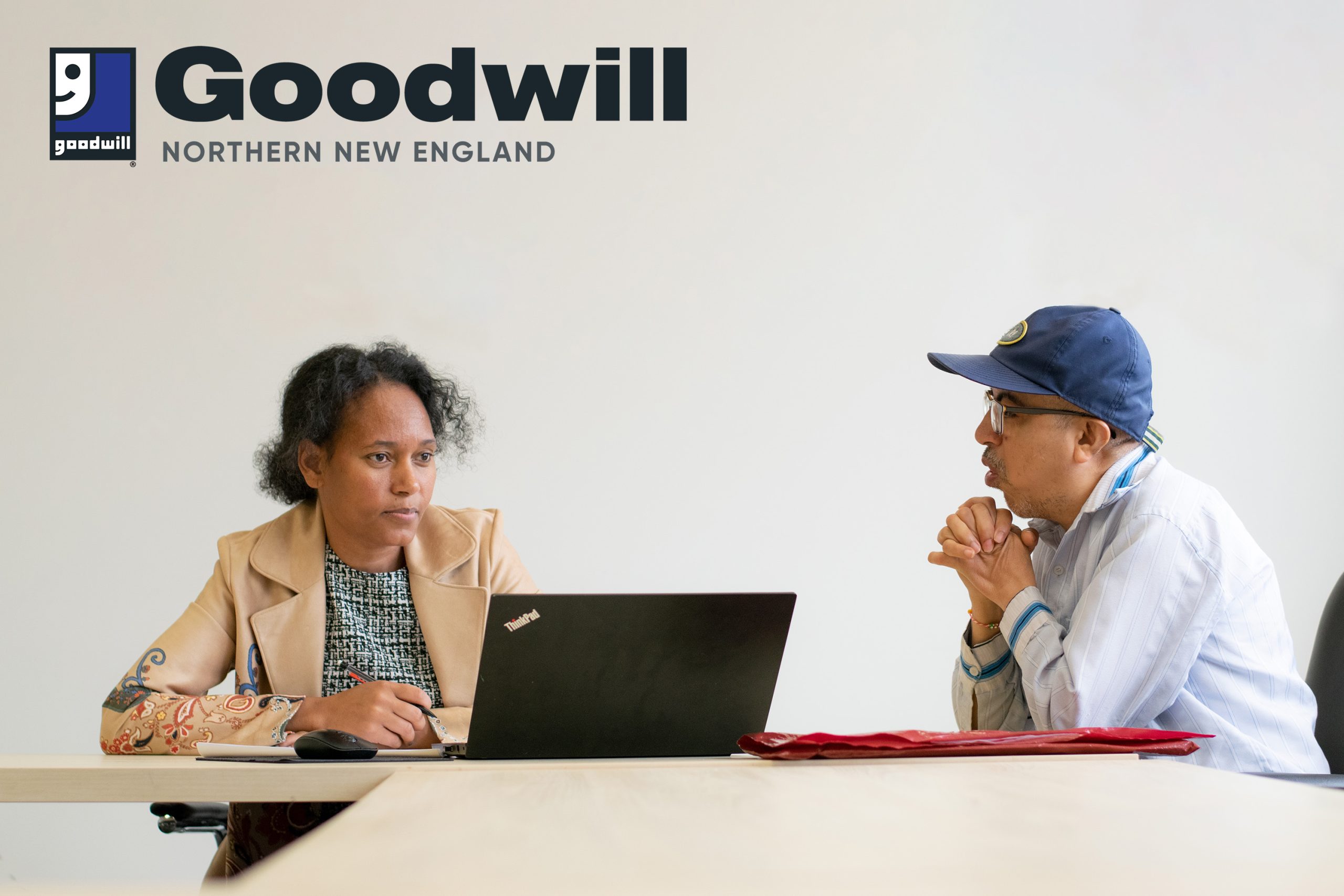 A woman in a blazer looks at a laptop while she talks to a man in a baseball camp with his head on his hands. She is a Life Navigator offering Life Navigation services from Goodwill