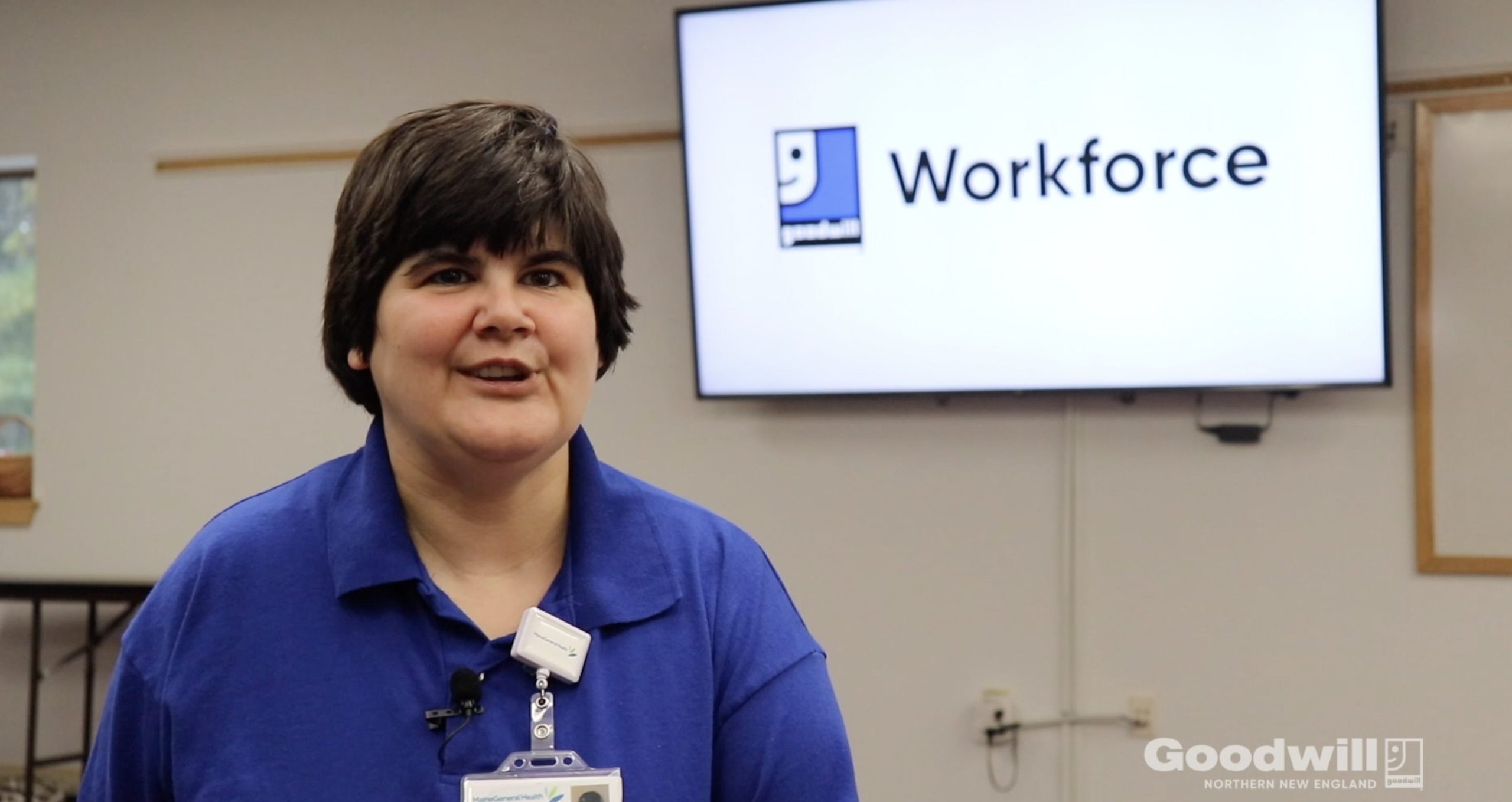 Angie stands in front of a Goodwill Workforce screen while at her job at Gray Birch in Augusta. She's wearing a blue shirt, and has black hair.