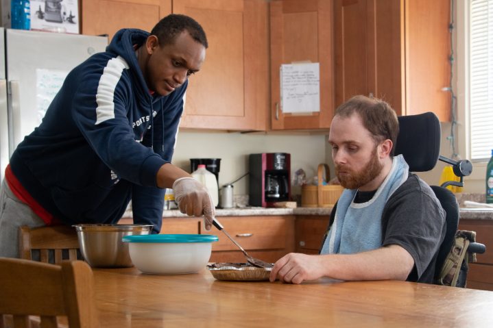 A Direct Support Professional DSP named Yannick is wearing kitchen gloves and helps a Goodwill Homes client Patrick smooth our a chocolate cream pie. Patrick is in a wheelchair and wears an apron as he watches. 