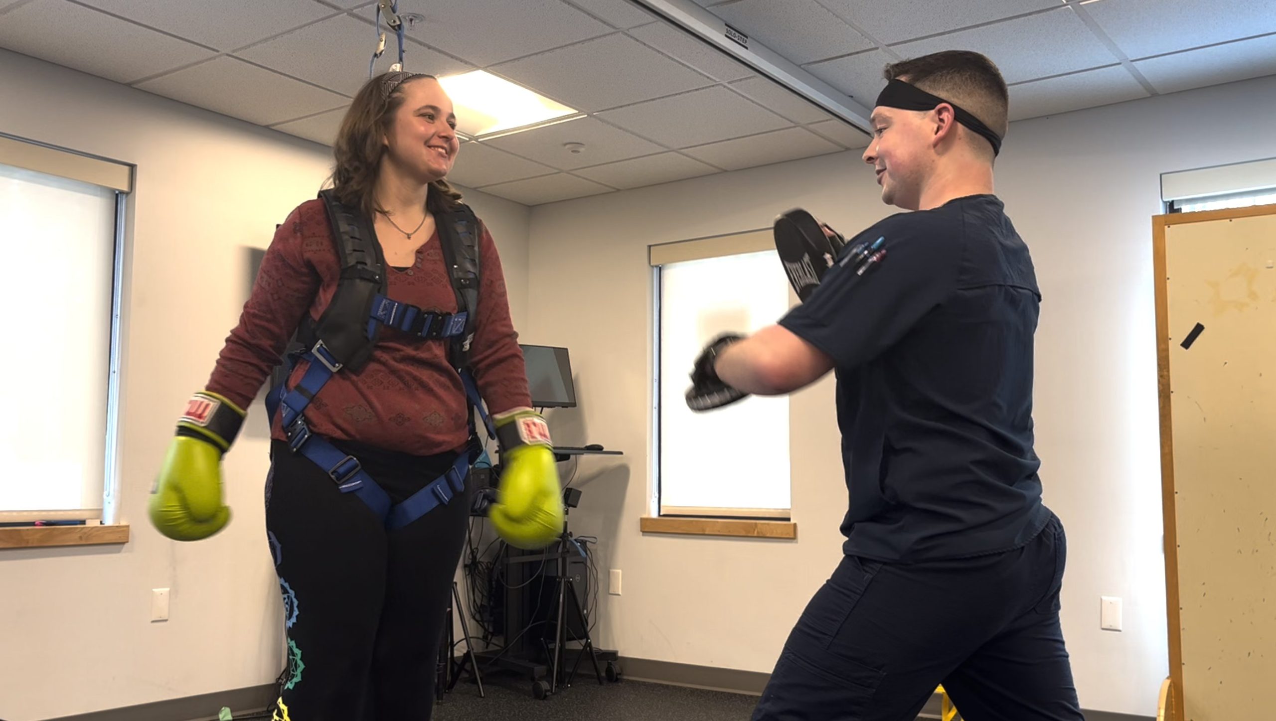 Emma is wearing a red shirt and yellow boxing gloves while hooked up to a ceiling harness. She is boxing with Kevin, a PT specialist at NeuroRehab.