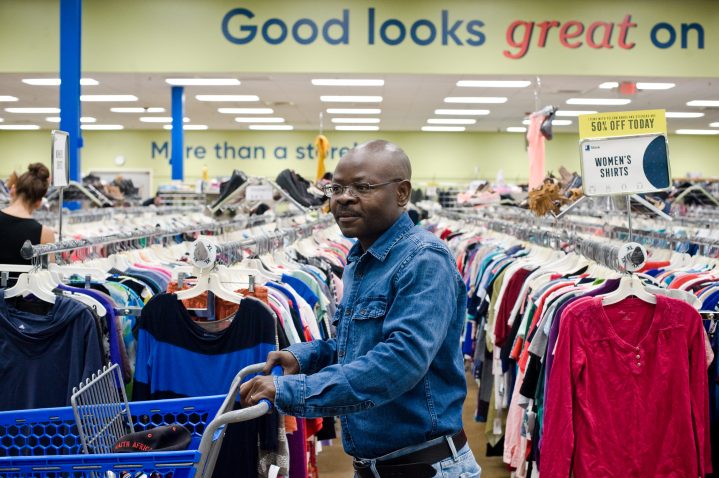 A man holds the handles of a blue shopping cart while browsing the aisles of clothing in a Goodwill store