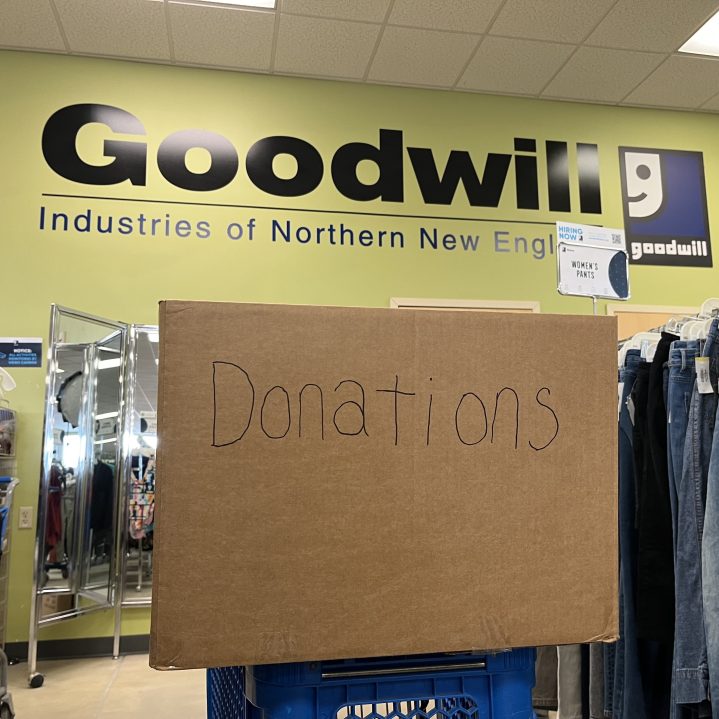 A cardboard box labeled "Donations" sits atop a blue shopping cart with Goodwill Northern New England's logo and name in large text on a green wall in the background.