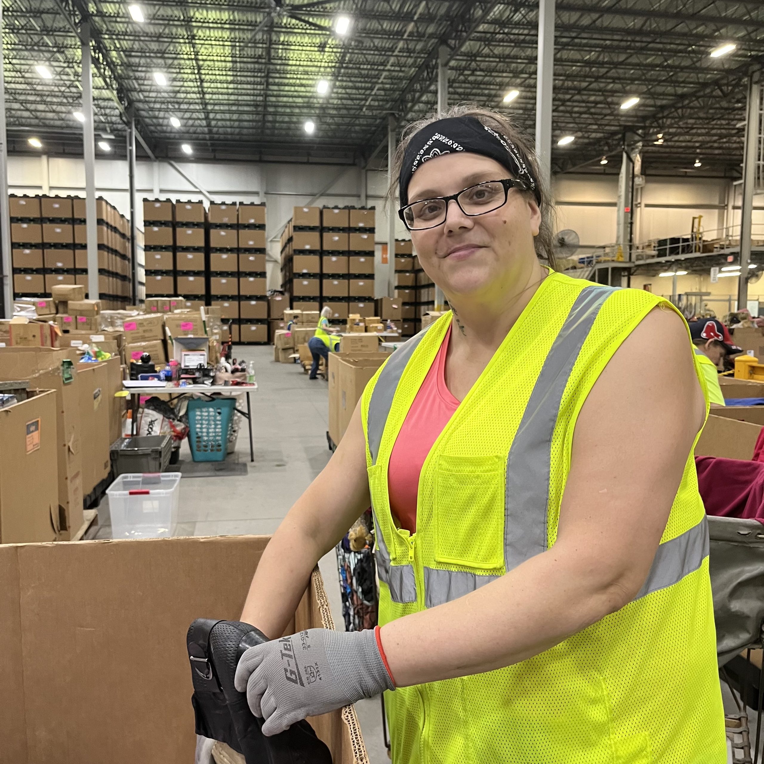 Amanda stands smiling next to a box of donations she is sorting. She is wearing a yellow high-visibility safety vest, a black headband and gray gloves. Behind her is a large stack of boxes containing donated goods.