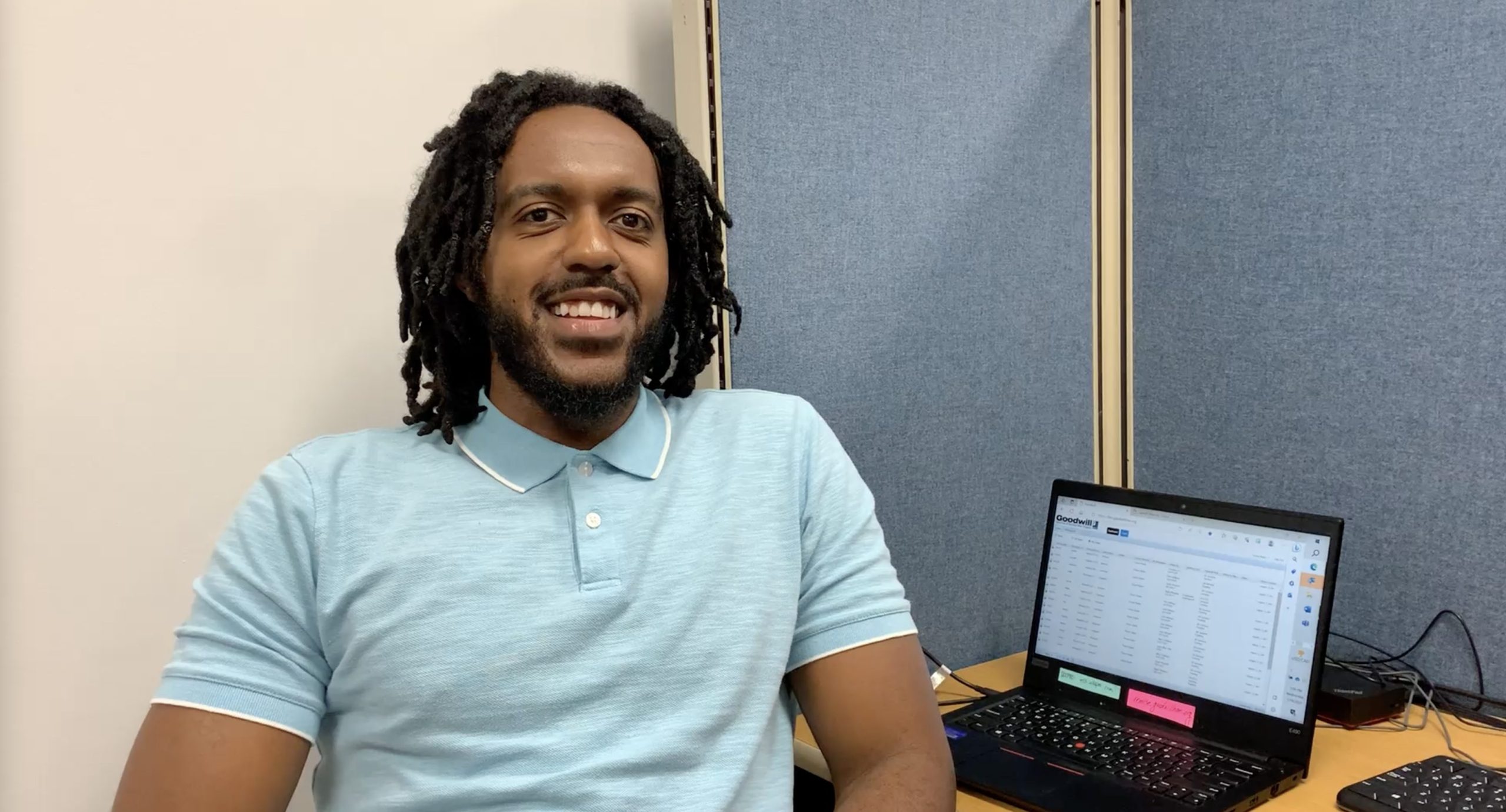 Trevon Butler is a career advisor or Goodwill. Here he sits, smiling, in a blue shirt against a white wall and blue cubicle. His laptop is in the background.