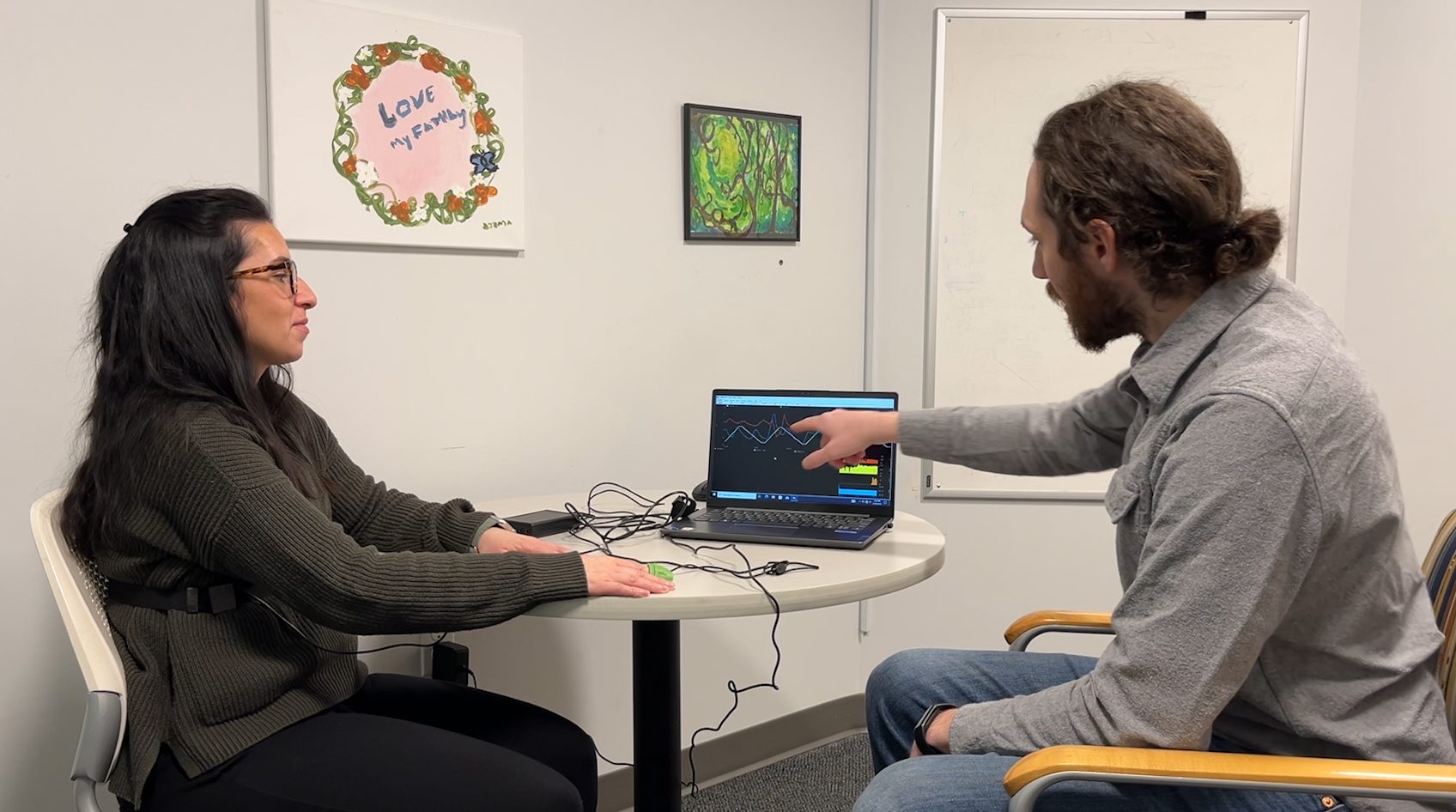 A woman is sitting at a table while wearing a heart rate monitor and respiratory monitor. A man points to a computer screen that shows the monitors' readings as a form of Biofeedback.