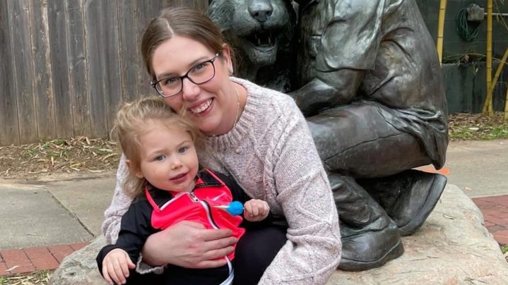 Victoria Sittig, holding her young daughter, and are sitting in front of a statue.They are smiling at the camera. 