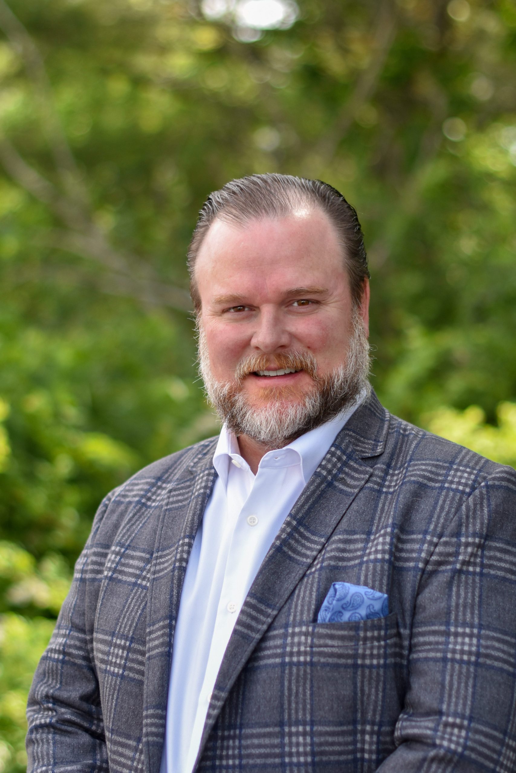 A man in a suit smiles while standing outdoors. There is greenery behind him.