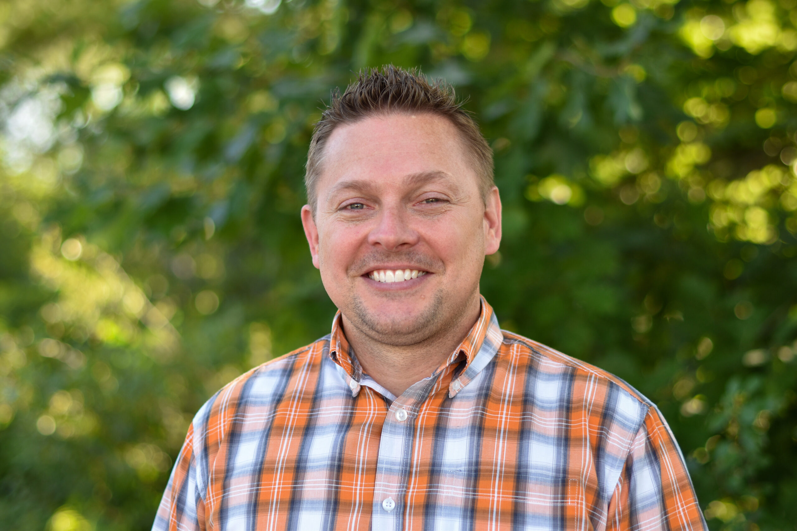 A man in an orange plaid shirt smiles outdoors. There is greenery behind him.