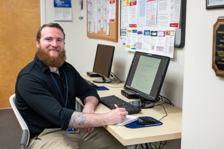 A man at a computer smiles and holds a pen to a notebook