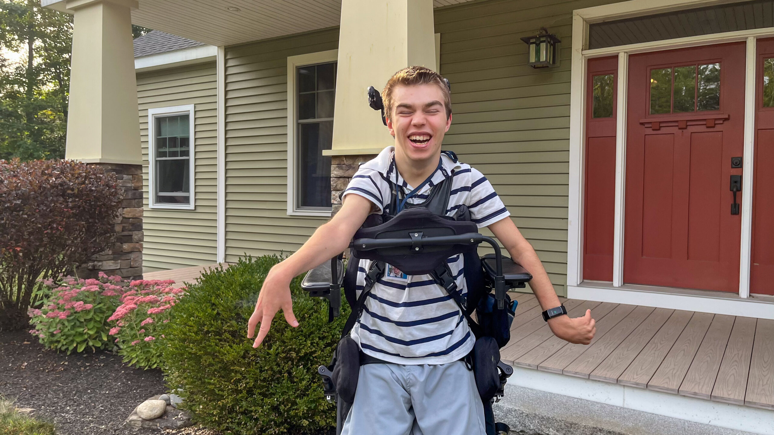 A young man in an upright wheelchair smiles outside a house.