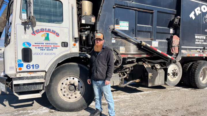 A man with a beard wearing a hat standing in front of a garbage truck.