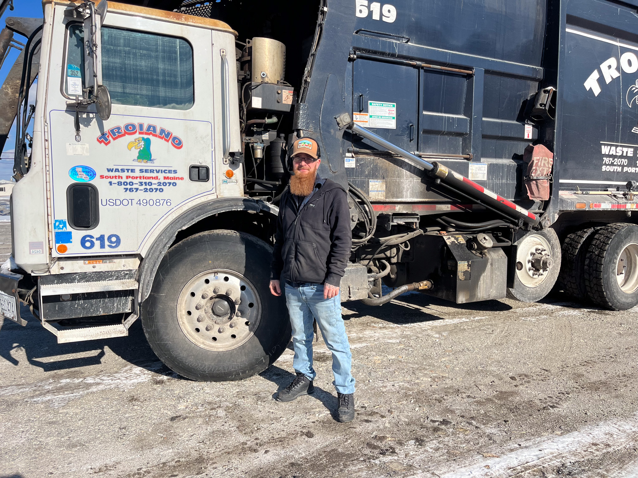 A man with a beard wearing a hat standing in front of a garbage truck