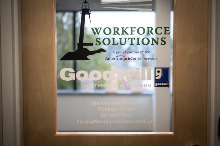 A wooden door with a glass pane. On the glass is a Goodwill Northern New England logo and Workforce Solutions logo.